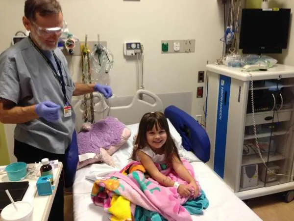 A hospital technician placing EEG leads while a young Brooke sits up in bed with a stuffed animal beside her.
