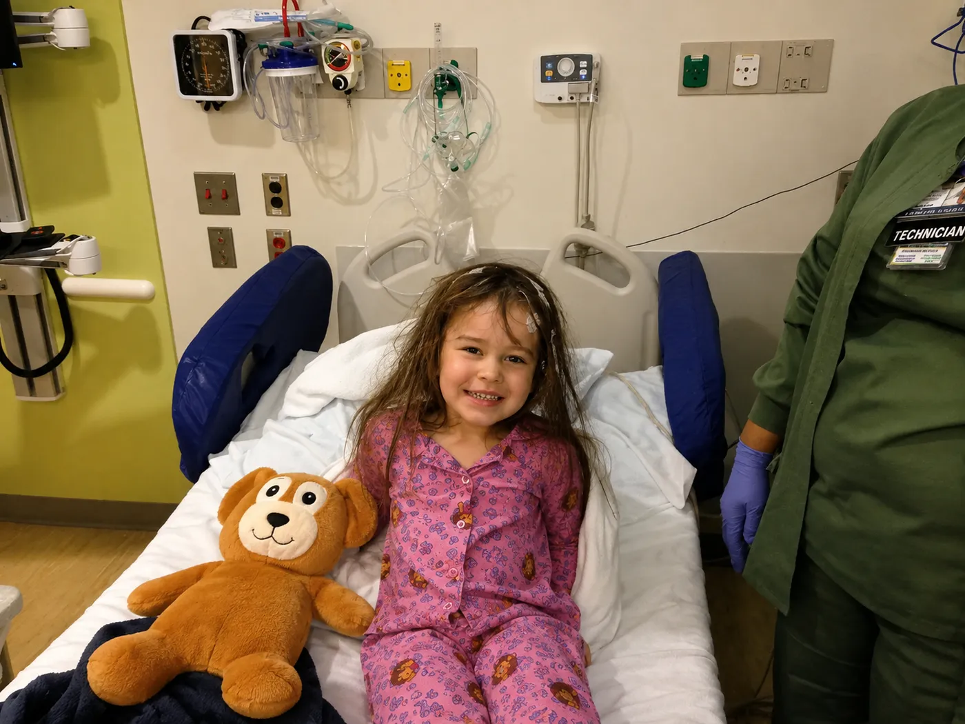 Three-year-old Brooke smiling in a hospital bed beside her new stuffed Seizure Bear, EEG leads still in her hair, as a technician stands nearby.