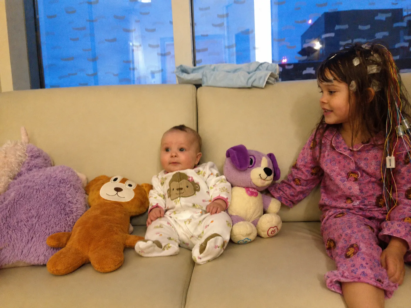 Brooke, wired for EEG monitoring, sitting on a hospital couch with her baby sibling and Seizure Bear lined up beside them.