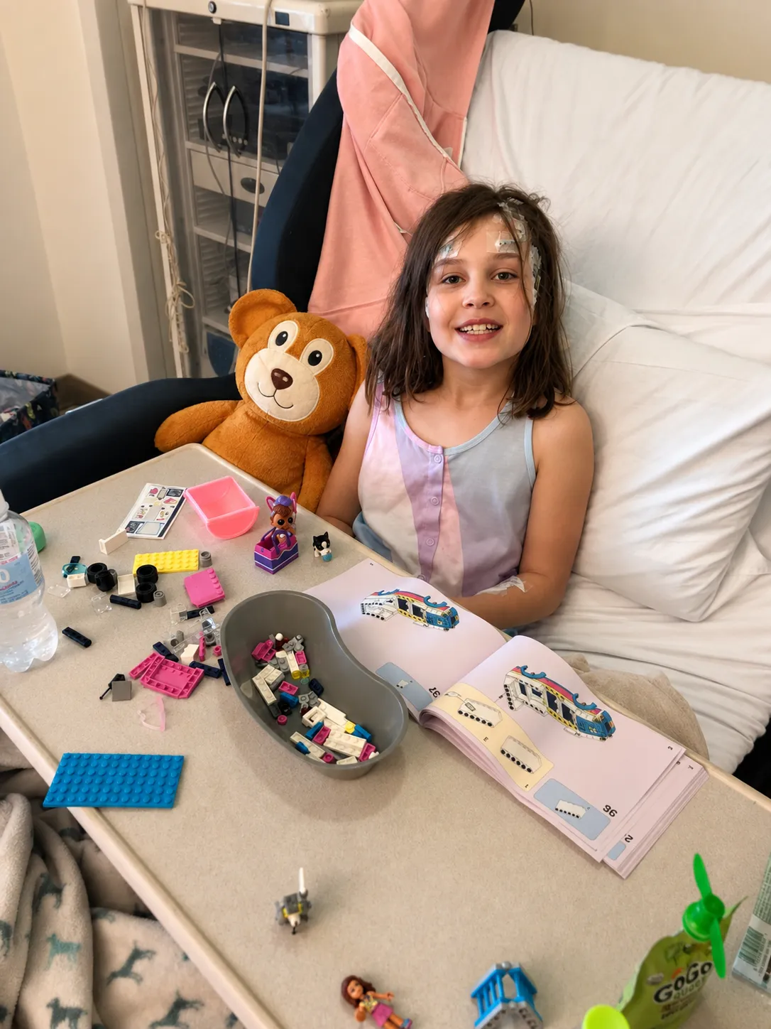 An older Brooke in a hospital bed with monitoring leads taped to her head, building a LEGO set at her tray table with Seizure Bear propped up beside her.